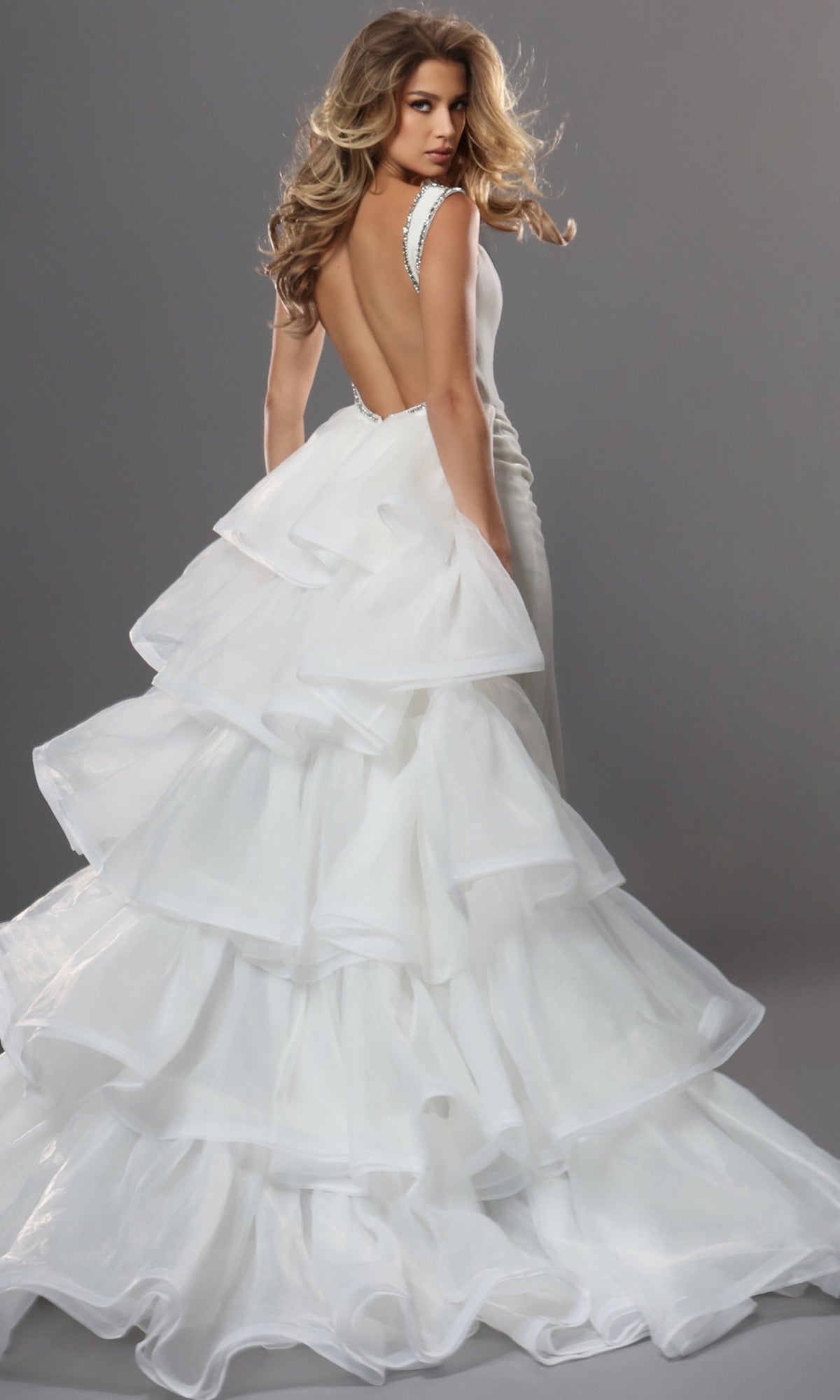 A woman wearing the Jovani 42661 Long Formal Dress, a backless white gown featuring a layered ruffle skirt, stands against a plain gray background and looks over her shoulder.